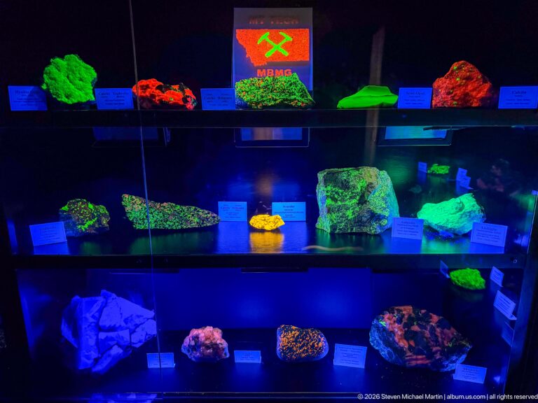 Wide dark display case filled with multicolored fluorescent minerals beneath an illuminated mining emblem.