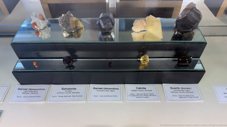 Two-tier museum case containing labeled Montana garnet, sphalerite, calcite, and smoky quartz specimens.