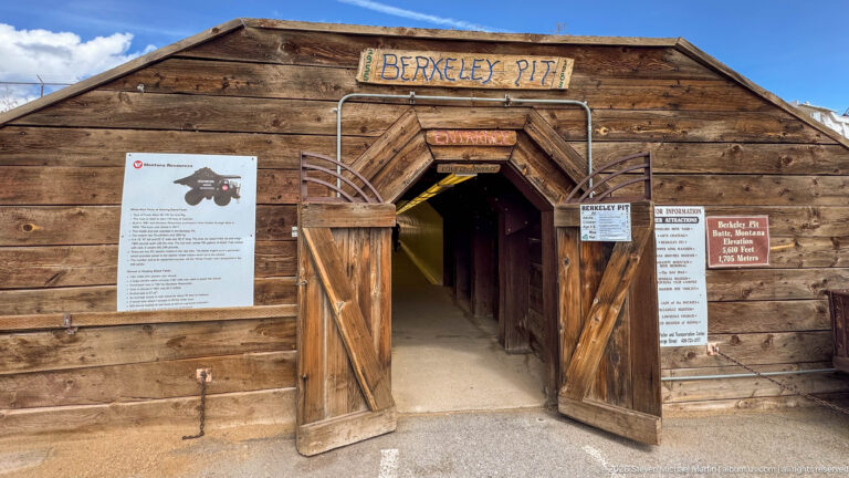 Centered timber Berkeley Pit entrance with open plank doors, angular portal framing, visitor signs, and dark passage.