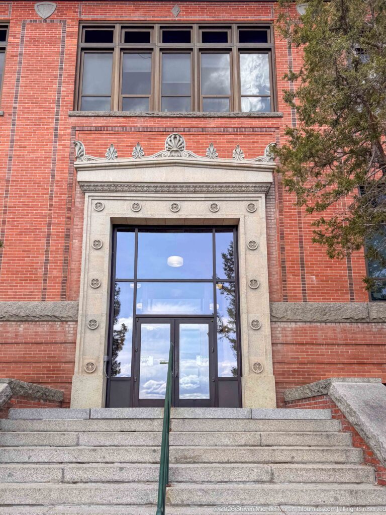 Stone steps leading to glass doors framed by carved pale stone in a red-brick historic institutional façade.
