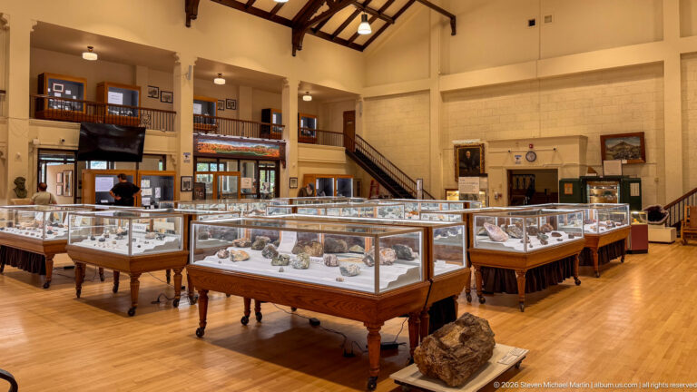 High-ceilinged museum gallery with rows of glass-topped wooden mineral cases, balcony, stairs, and framed displays.