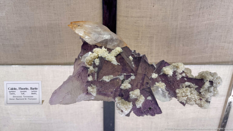 Large clear calcite crystal above purple fluorite and pale clustered minerals mounted in a display case.