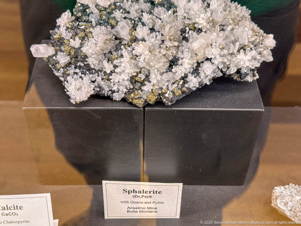 Dark sphalerite specimen with pale quartz crystals and metallic pyrite displayed in a mineral case.