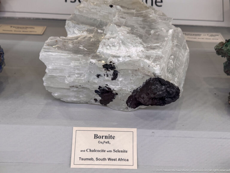 Clear to white crystalline mineral mass with dark bornite patches, displayed above an identifying museum label.