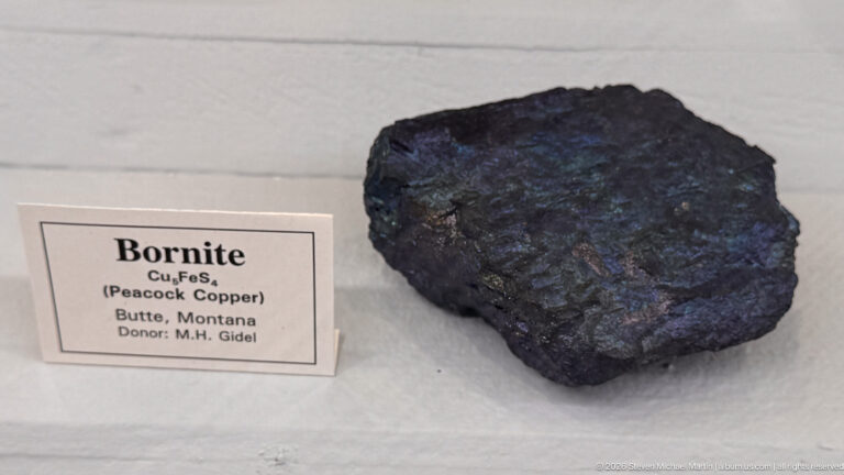 Dark metallic bornite mineral specimen beside a label identifying Cu5FeS4 and Butte, Montana.