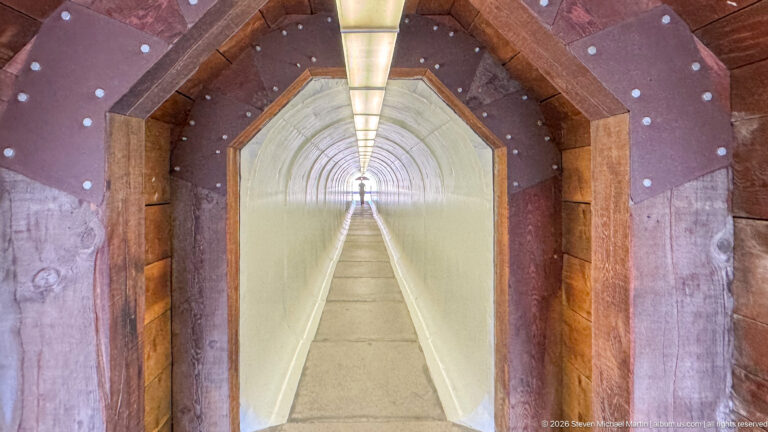 Long reconstructed mine tunnel with repeated timber supports, pale walls, central walkway, and continuous overhead lighting.