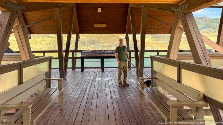 Covered timber viewing deck with benches, a visitor and dog, and Berkeley Pit water and terraced walls beyond.