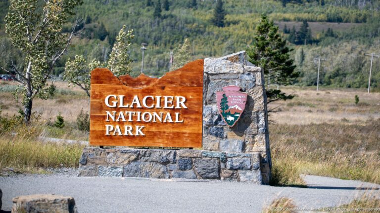 Wood-and-stone Glacier National Park sign with National Park Service arrowhead beside a grassy roadside.