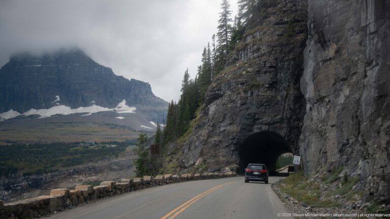 Car approaches a small tunnel portal cut through a cliff beside stone guardwalls and clouded mountains.