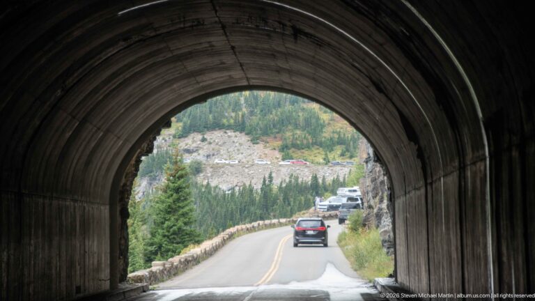 Cars travel beyond the rounded dark interior of a short tunnel on Going-to-the-Sun Road.