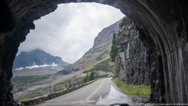 Going-to-the-Sun Road curves from a dark rock tunnel toward mountains and lingering snow.