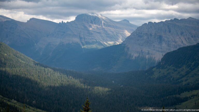 Deep forested mountain valley framed by rugged peaks with sunlight on a distant slope.