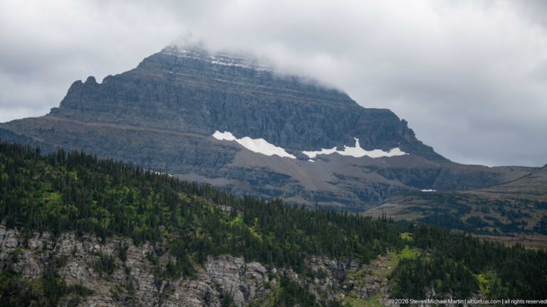 Layered mountain with lingering snow patches rises above conifer forest into low cloud.
