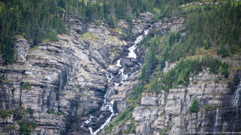 Multi-tier white waterfall drops through a steep gray rock gorge bordered by conifers.