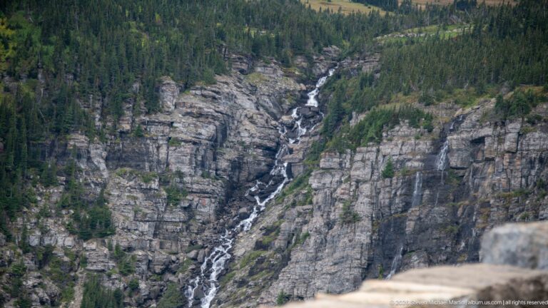 Long waterfall descends a layered rock gorge between dense conifer forests.