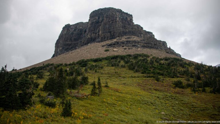 Blocky layered mountain summit rises above an alpine meadow with scattered conifers.