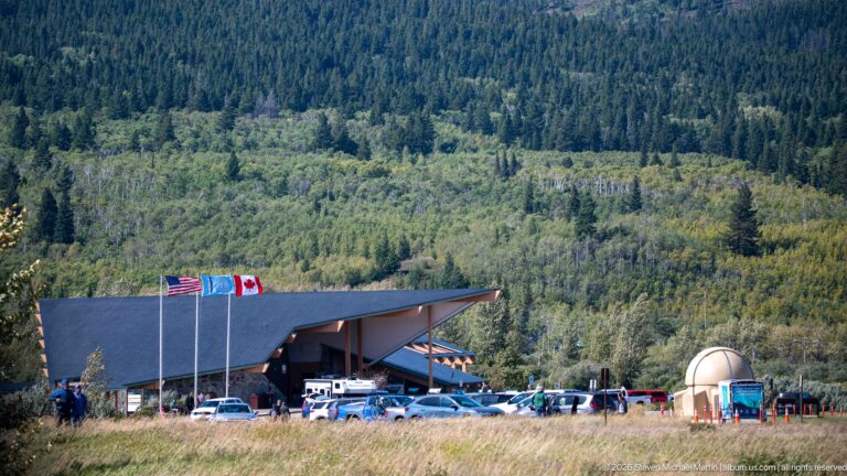 Angular-roofed park facility with parked cars and United States and Canadian flags before a forested hillside.