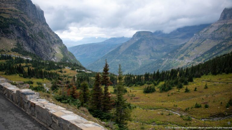 Stone road barrier overlooks a green alpine valley enclosed by steep clouded mountains.