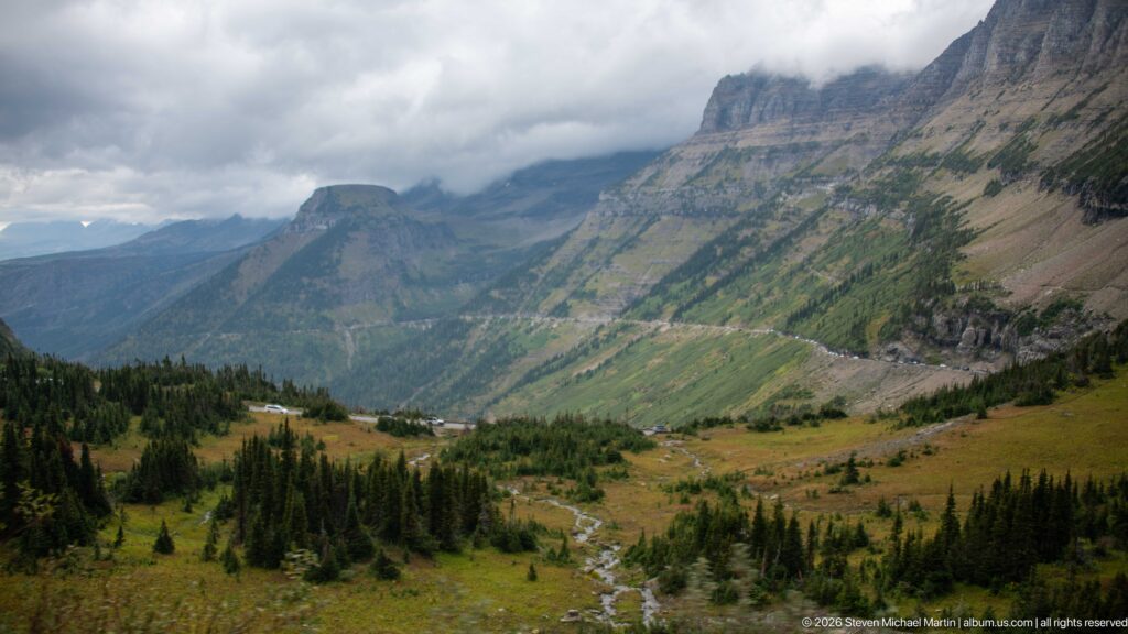Alpine valley with winding stream, scattered conifers, steep mountains, and road beneath low clouds.