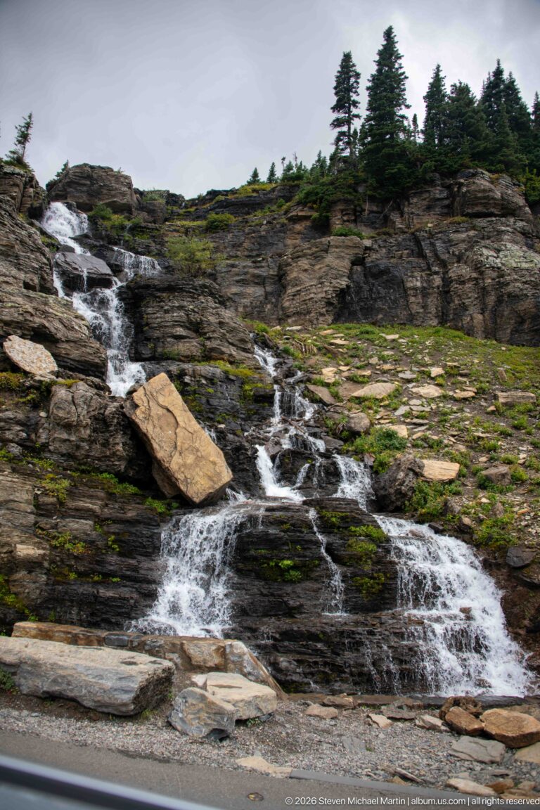 White cascade spreads across dark bedrock around a large tan boulder beside the road.