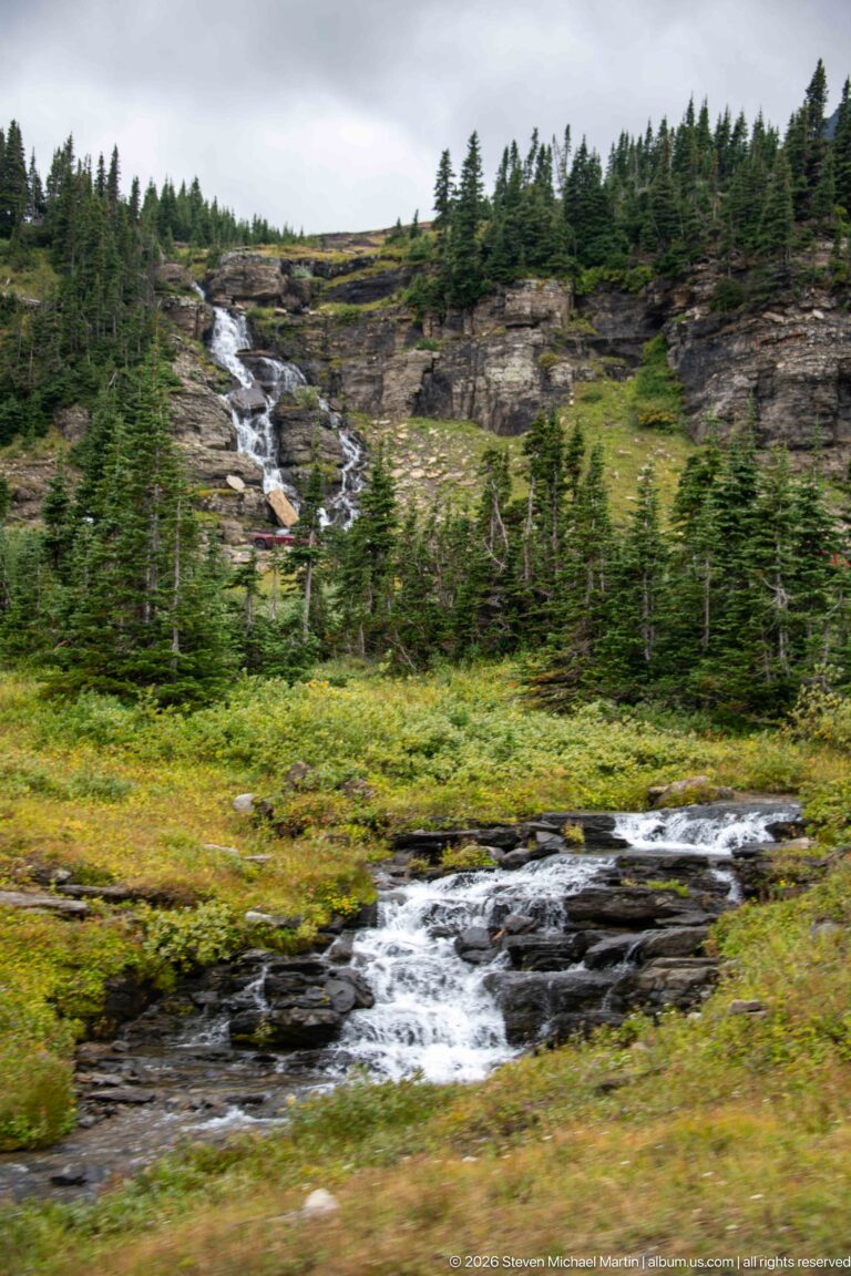 Alpine stream cascades over dark rocks through green meadow with conifers and a second cascade behind.