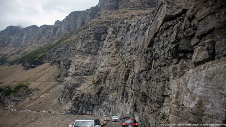 Huge stratified cliff rises above a line of vehicles on the narrow mountain road.