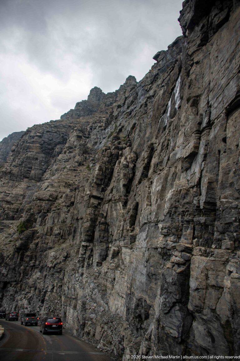 Layered gray cliff towers directly above cars traveling Going-to-the-Sun Road.