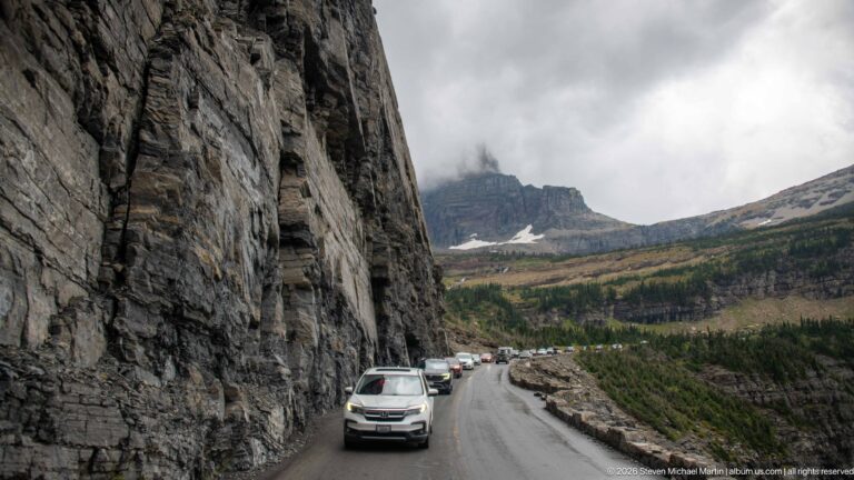 Vehicles travel wet mountain pavement beside a sheer cliff with a clouded peak ahead.