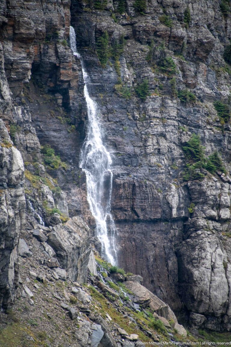 Narrow white waterfall descends a layered gray rock face dotted with small conifers.
