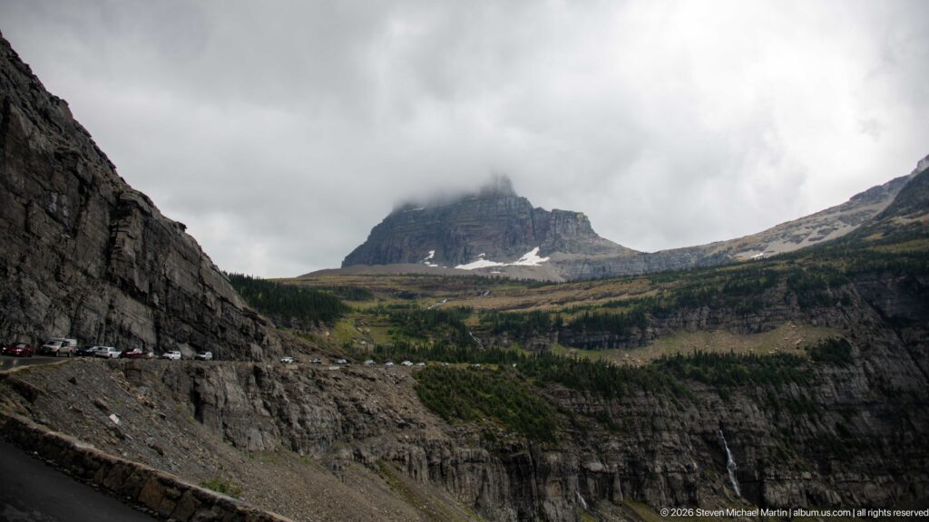 Cars follow Going-to-the-Sun Road below towering layered cliffs and a cloud-covered peak.