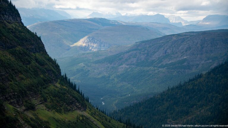Deep forested mountain valley with layered rock walls and distant peaks beneath cloudy sky.