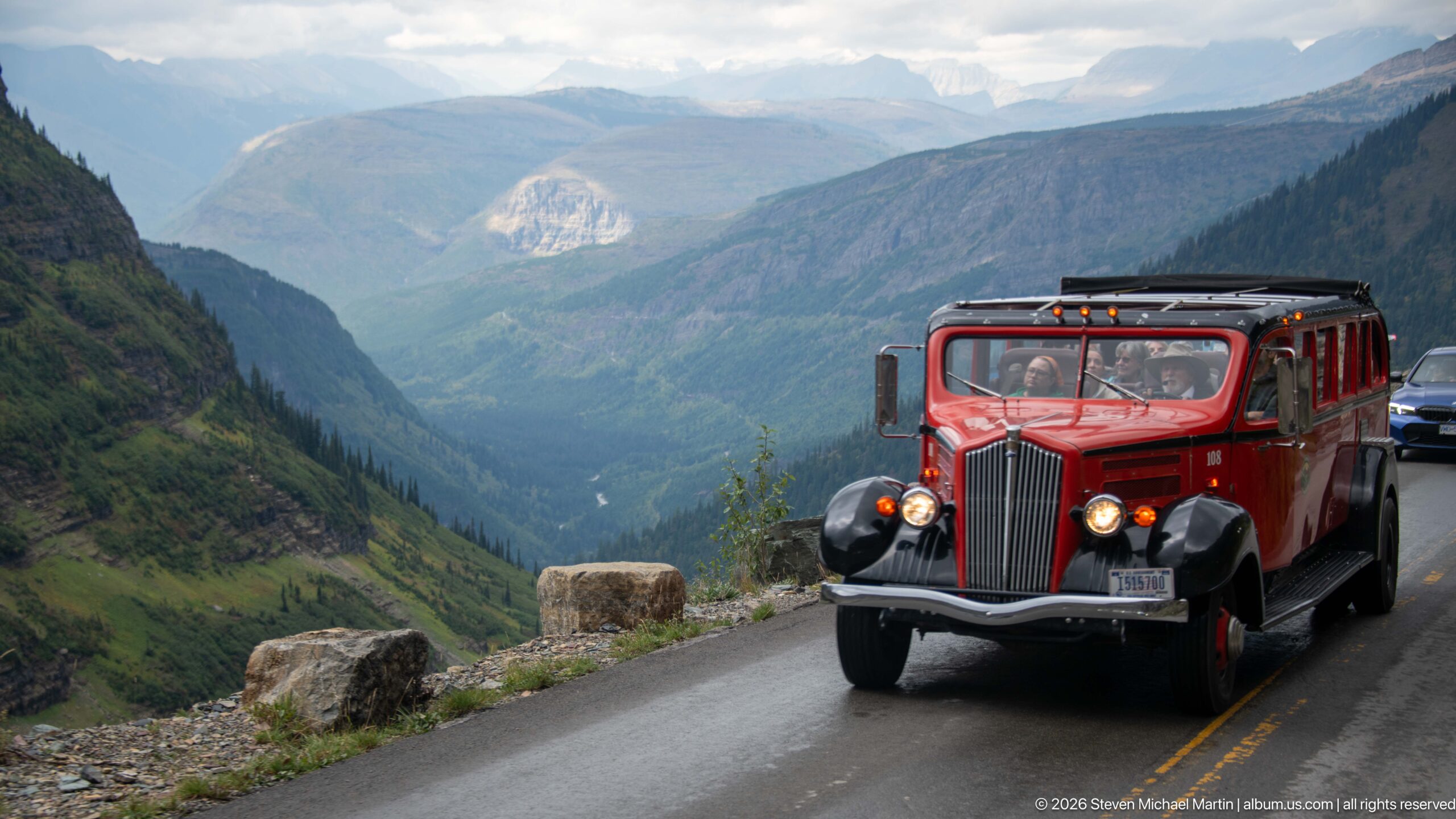 Red vintage-style tour bus travels a narrow mountain road above a deep forested valley.