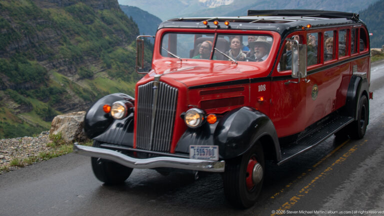 Close view of a bright red vintage-style Glacier tour bus carrying passengers on a mountain road.