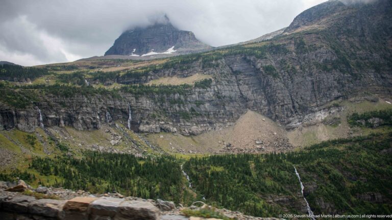 Layered mountain cliffs above a green basin with narrow waterfalls, a pale talus fan, snow patches, and low clouds.