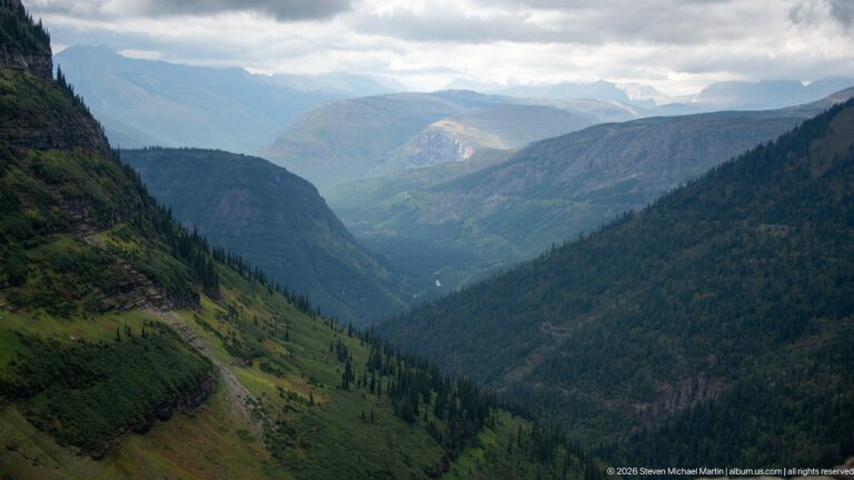 Deep mountain valley extending toward distant ridges, with steep forested slopes and open drainage corridors beneath broken clouds.