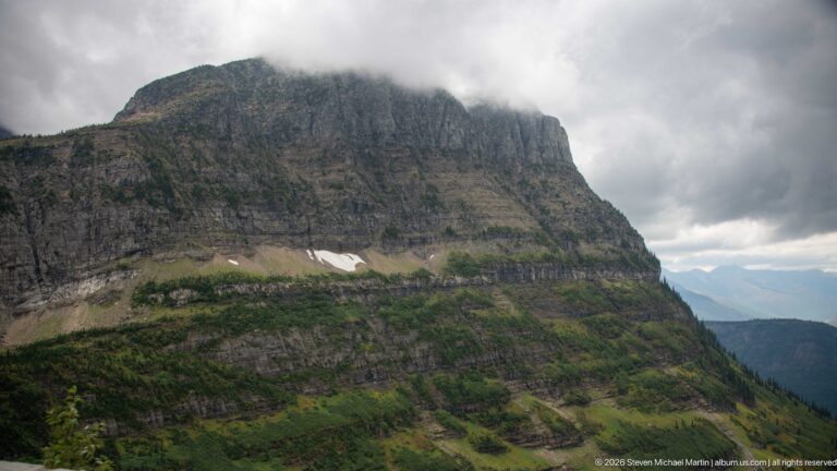 Massive layered mountain cliff rising above green slopes and small snow patches, with its summit hidden by low clouds.