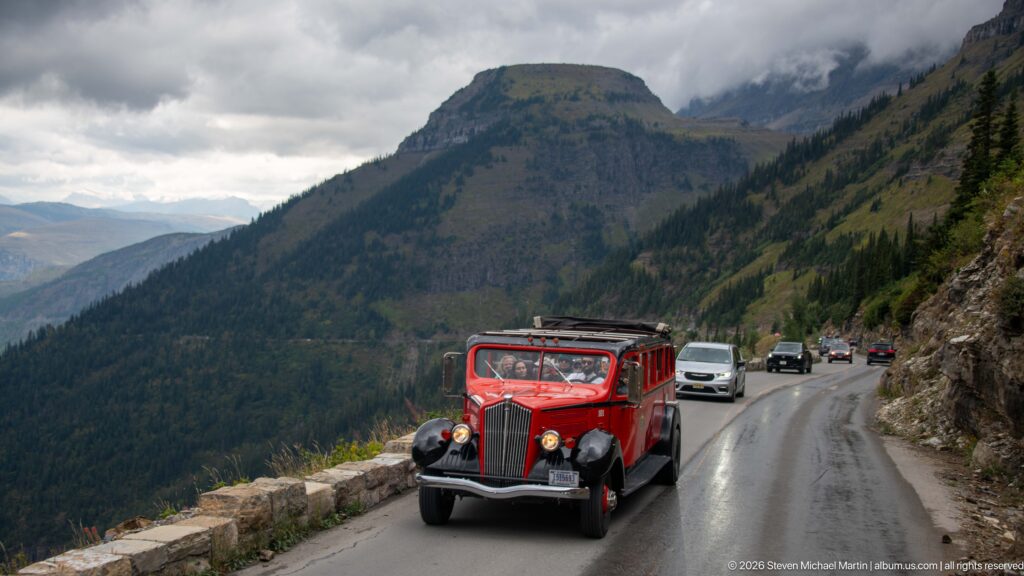 Bright red Glacier tour bus approaching on a narrow cliffside road beside a stone guardwall and other vehicles.