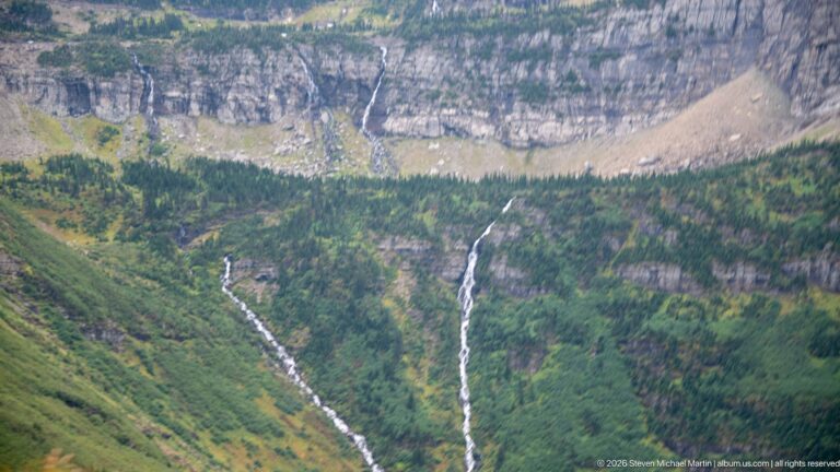 Two long white waterfalls descending a green mountainside below a massive layered cliff and pale talus slope.