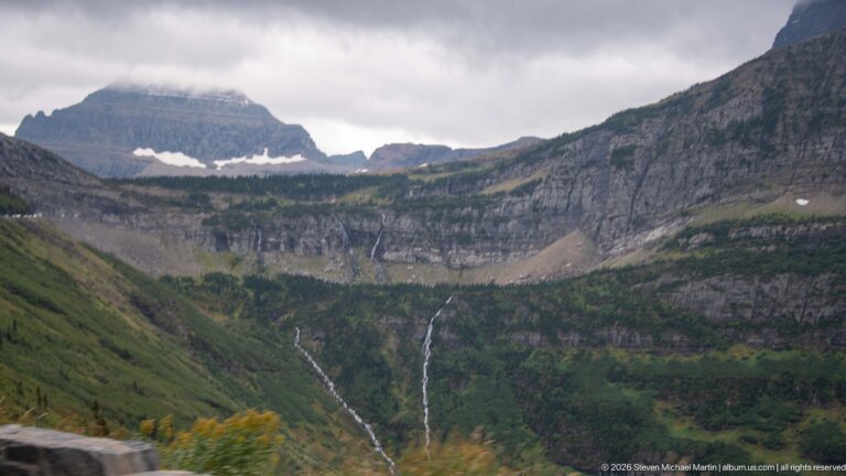 Two narrow waterfalls dropping from a high rocky basin beneath layered cliffs and lingering snow patches.