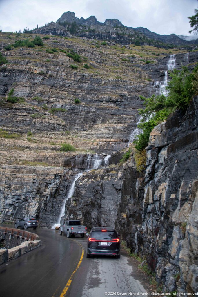 Cars on wet Going-to-the-Sun Road beneath a broad multi-channel cascade descending a huge stepped rock face.
