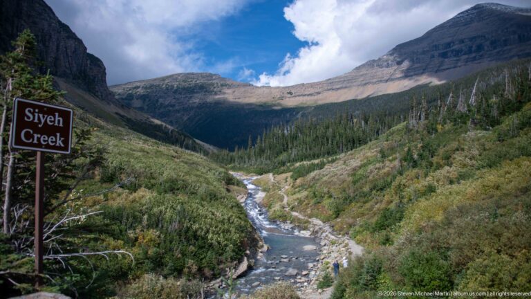 Siyeh Creek sign stands beside a rocky creek flowing through a steep forested mountain valley.