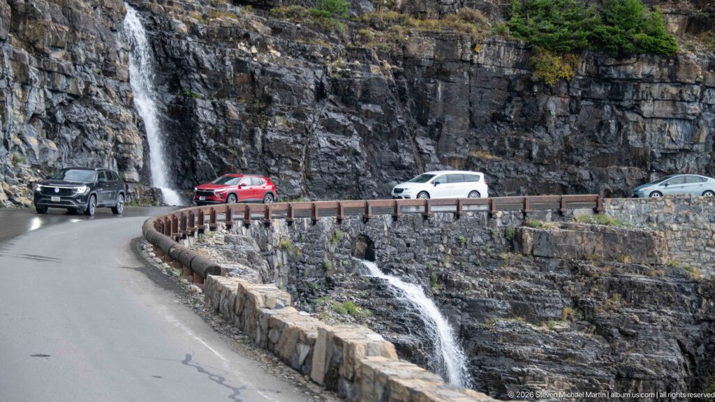 Cars rounding a narrow mountain-road curve beside a waterfall, stone retaining wall, and dark layered cliff.