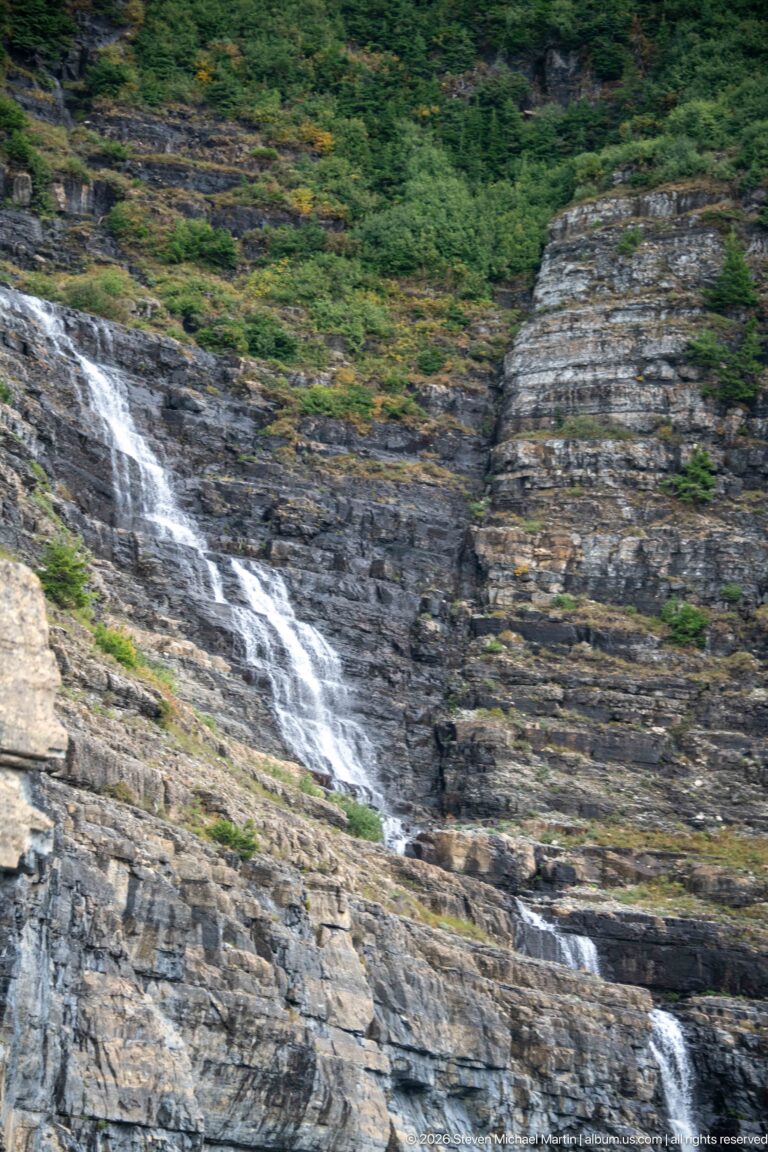 White cascade descending diagonally across layered gray-brown bedrock below dense green shrubs.