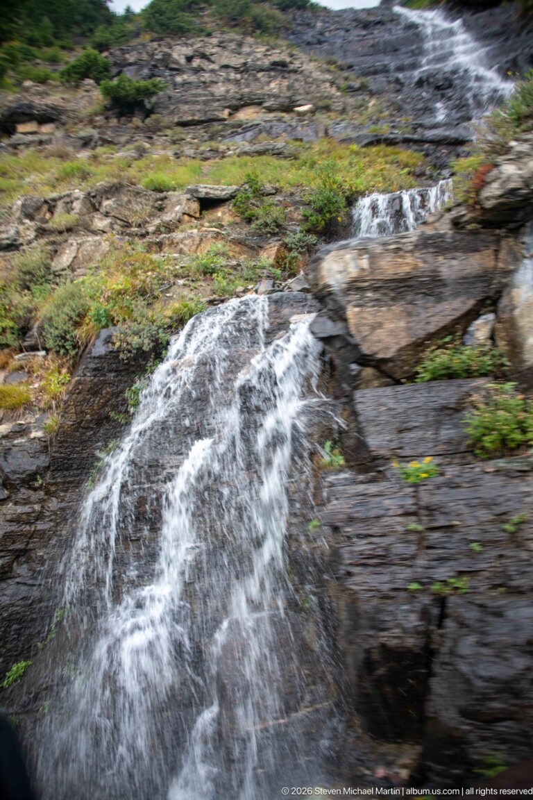 White mountain runoff rushing over dark fractured rock ledges bordered by low green vegetation.