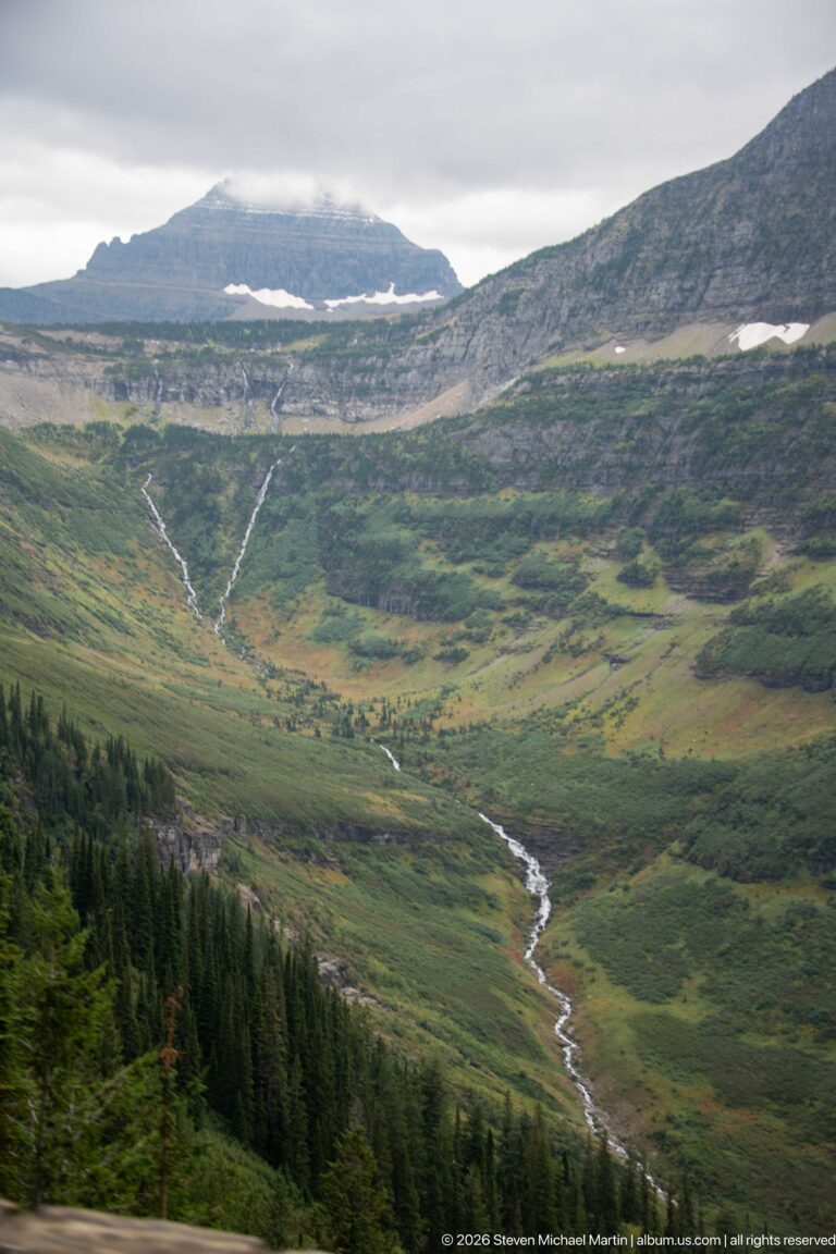 Winding alpine stream crossing a broad green valley beneath layered cliffs and lingering snow patches.