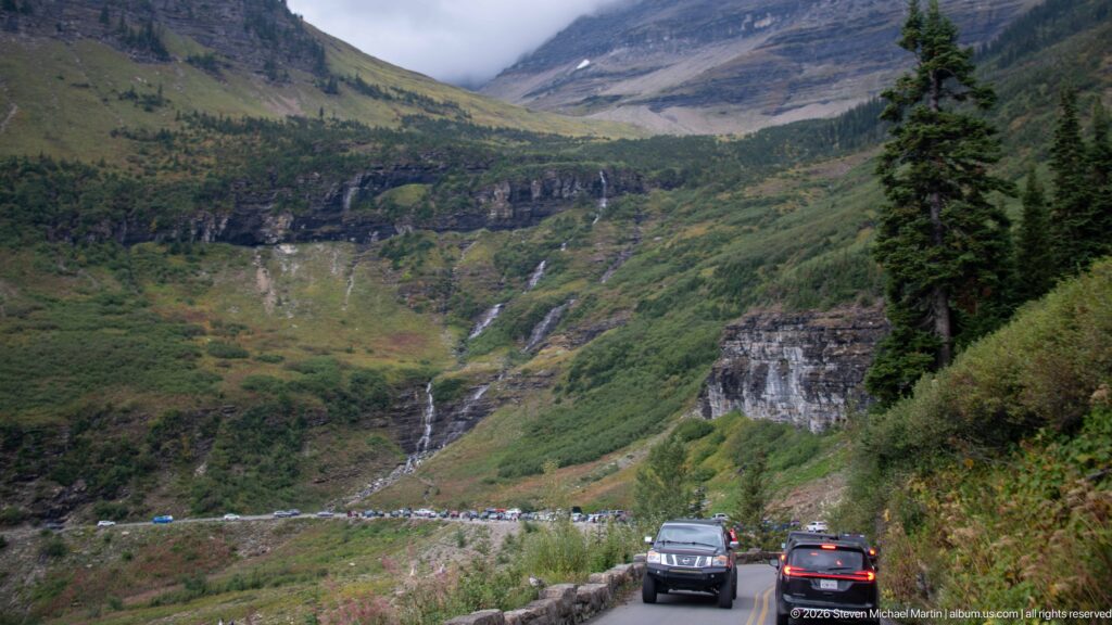 Cars traveling a narrow mountain road toward a bend below green cliffs and a slender descending waterfall.