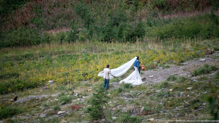 Bride in a white gown on a rocky mountain slope while an assistant spreads the long train and another person stands nearby.