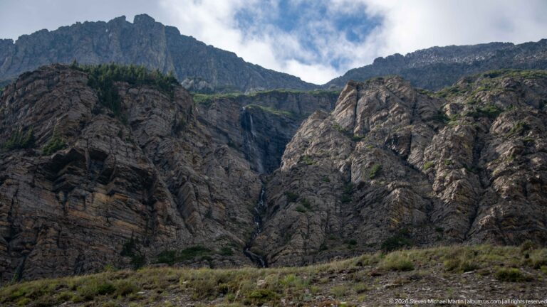 Thin waterfall descends a dark ravine between massive rugged rock walls beneath blue sky.