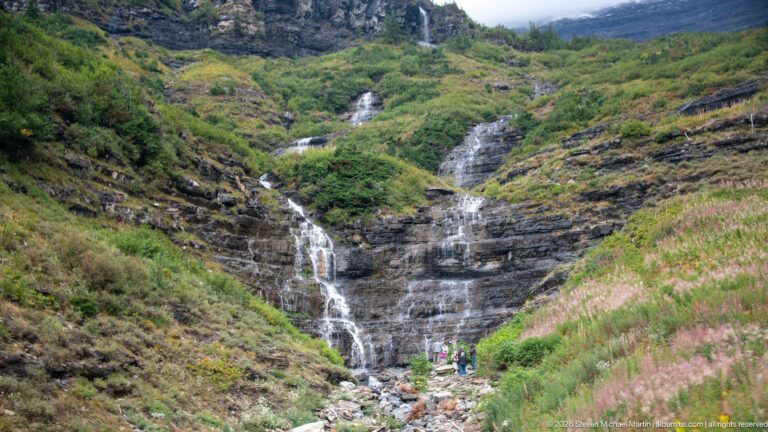 Multi-tier waterfall descending dark rock ledges through green vegetation, with several people standing near the rocky base.