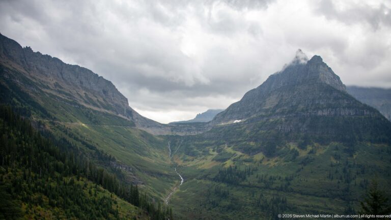 Sharp layered mountain peak rising above a green valley and narrow stream beneath thick gray clouds.